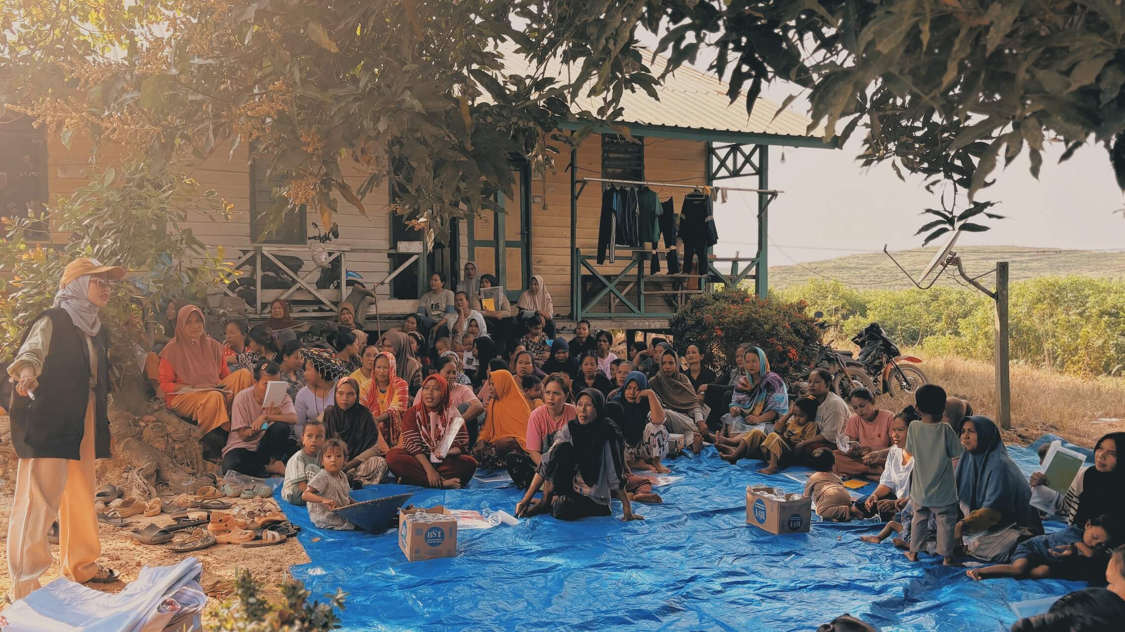 Community members gather on a blue tarp beneath a wooden village house, with fields visible behind them.