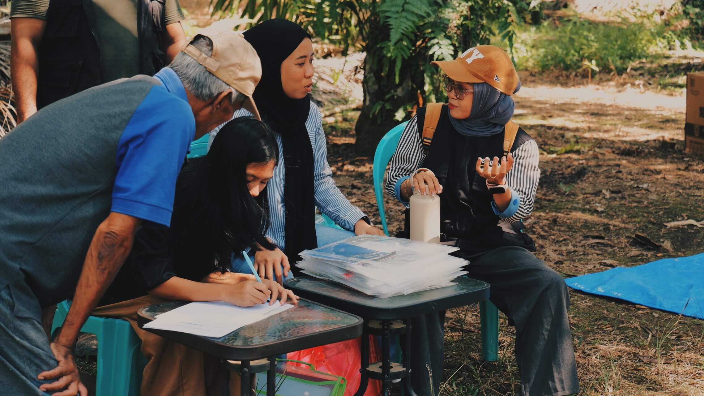 A small group works around tables outdoors, discussing notes and documents under the trees.